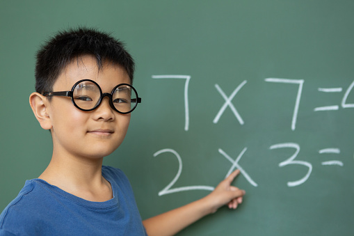 Front view of a smart Asian schoolboy doing math on greenboard in a classroom at elementary school