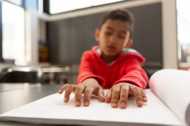 Front view of blind mixed-race schoolboy hands reading a braille book at desk in a classroom at elementary school