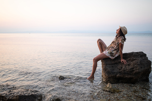 Young  woman enjoying alone by the sea on sunset, sitting on rock with feet in the water