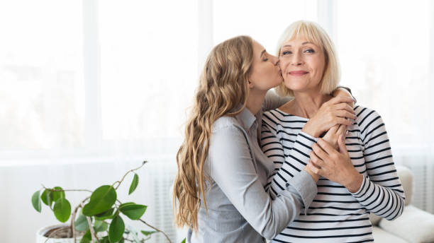 Young daughter kissing senior mother on the cheek Young daughter kissing senior mother on the cheek, standing against window at home, copy space cheek stock pictures, royalty-free photos & images