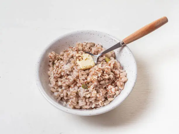 Photo of Barley porridge with a piece of butter, pumpkin and sunflower seeds, sesame seeds in a deep light ceramic plate, a teaspoon with a wooden handle on a white background is next to it, removed from close distance