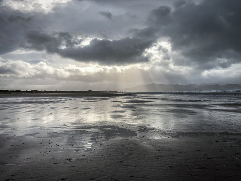 Moody Shot of a Beach at Low Tide on a Cloudy, Stormy Day - with Reflective Sheets of Water Below and Rays of Sun Shining Through the Clouds Above