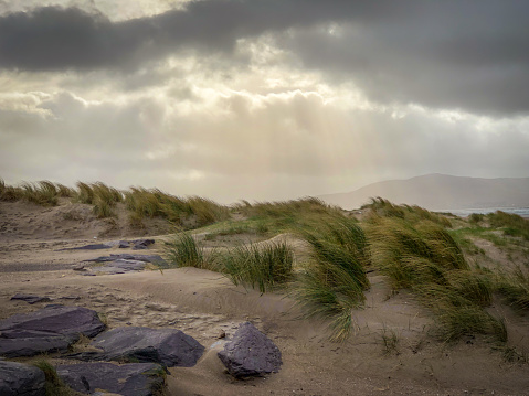 Moody Shot of Sand Dunes with Rocks and Patches of Grass in Front on a Cloudy, Overcast Day - with Rays of Sun Shining Through the Clouds Above