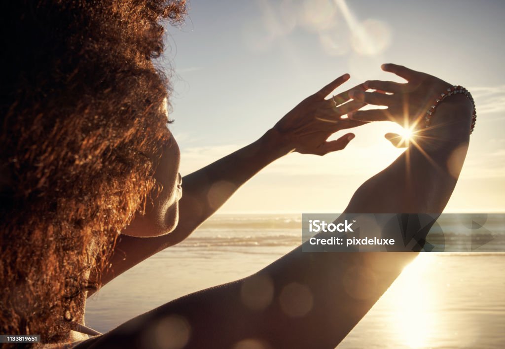 The ocean breeze brings a life of ease Rearview shot of a young woman blocking the sunlight with her hands at the beach Sunlight Stock Photo