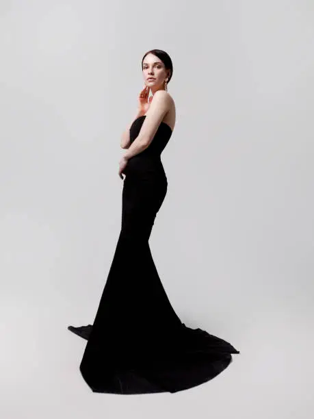 Full length portrait of young beautiful brunette woman wearing luxury strapless long black dress with train on white background. Designer clothing evening gown