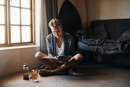 Shot of a young man reading a book while sitting on the floor at home