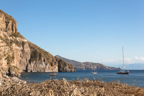 A serene view of Lipari Island's rocky coastline set above the clear, blue Mediterranean waters. The natural landscape features distinct vegetation and coastal rock formations against a calm and inviting sea.