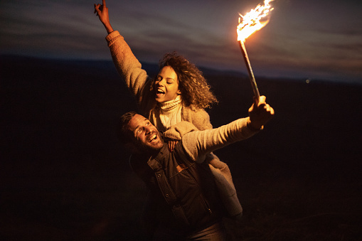 Young happy couple piggybacking and having fun with flaming torch by night.