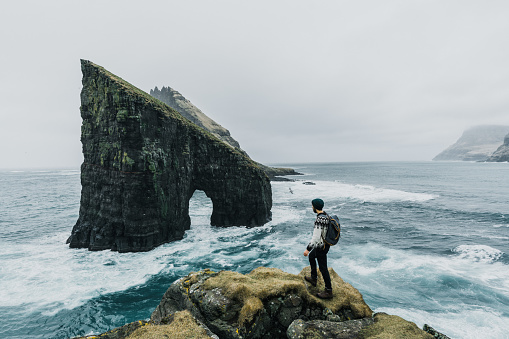 Young Caucasian man  in knitted sweater looking at view of Drangarnir arch in Faroe Islands