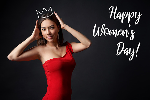 Beautiful woman in red dress supporting her crown with congratulation text of March, 8 International Women's day, over dark background