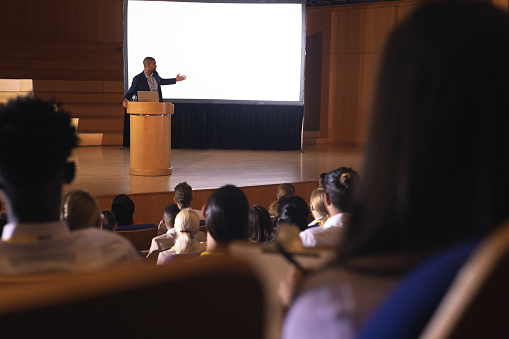 Rear view of audience sitting and listening while mixed race businessman standing near podium and giving presentation on white projector in the auditorium