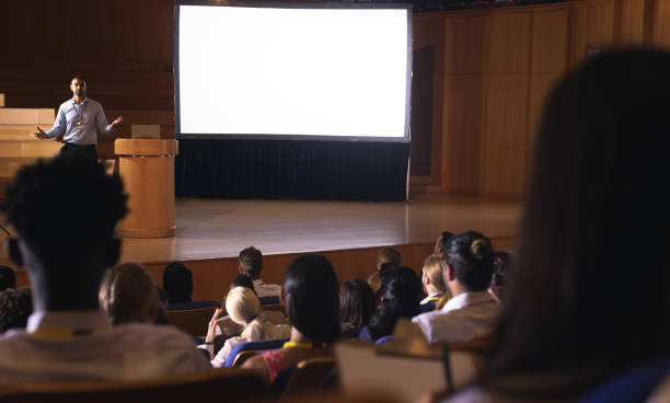 Rear view of mixed race audience listening to the presentation while mixed race businessman giving presentation in auditorium