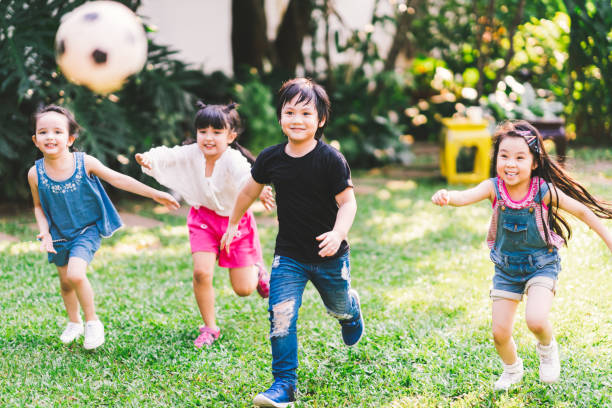 Asian and mixed race happy young kids running playing football together in garden. Multi-ethnic children group, outdoor sport exercising, leisure game activity, or childhood fun lifestyle concept stock photo