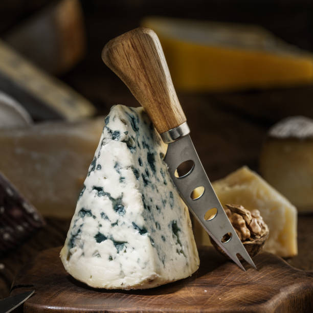 Segment of blue mould cheese and cheese knife on wooden board. Different cheeses at the background.