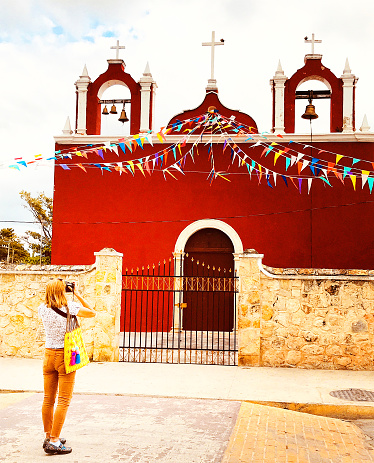 Yucatan, Mexico: An active senior female tourist taking a photo of a beautiful red Spanish Colonial Church in the Yucatan Peninsula. Shot in the coastal village of Celestún, about 60 miles from Merida.