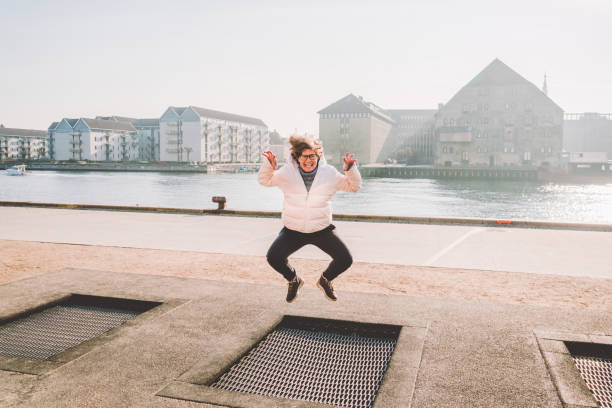 adult person rejoices like child. playground trampoline in ground, children trampoline, springs throws people up fun and cool. copenhagen river embankment denmark. woman jumping on street trampoline - women jumping bouncing spring imagens e fotografias de stock