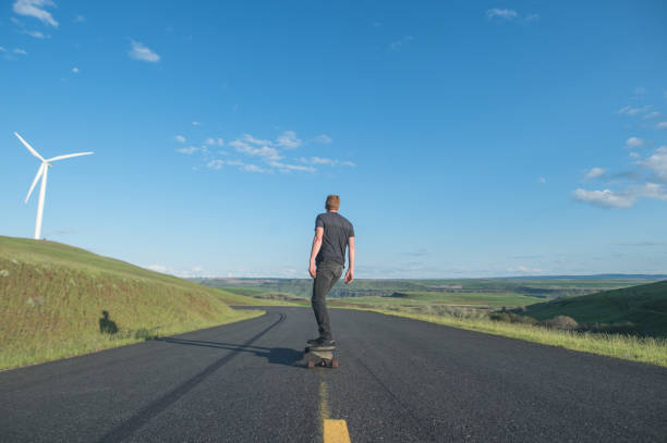 A man longboards down a rural highway surrounded by rolling hills and wind farms. The shot is from behind as he rides downhill.