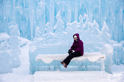 horizontal image of a caucasian woman on vacation sitting on an ice bench at the ice castles in the winter time.