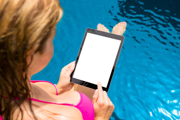 istock Woman by the pool holding tablet vertically with empty white screen. 1133938299