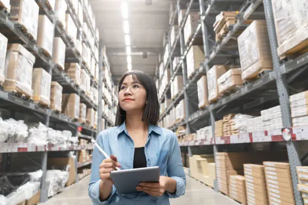Photo of Candid of young attractive asian woman auditor or trainee staff work looking up stocktaking inventory in warehouse store by computer tablet with wide angle view. Asian owner or small business concept.