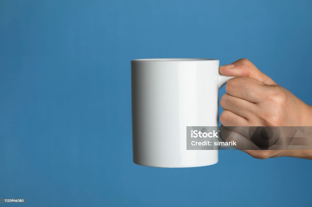 Relaxing Caucasian female is holding a white coffee mug in hand in front of a blue background. Mug Stock Photo