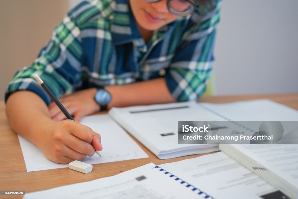 close up young asian student girl using pencil for writing formula of calculus on paper for do homework in library silent zone with textbook , campus lifestyle concept Educational Exam Stock Photo