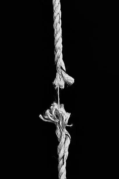 A rope hanging vertically against black is down to its last strand and about to snap. Monochrome image.