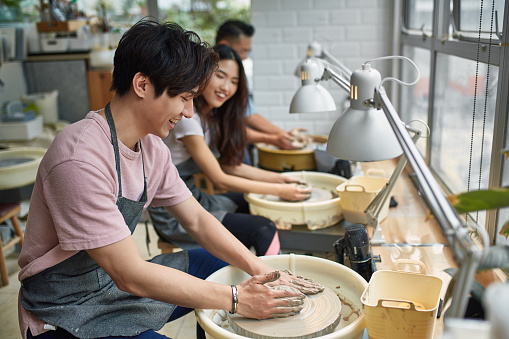 Trainees molding mud in pottery. Students are using spinning wheel in workshop. They are enjoying in training class.