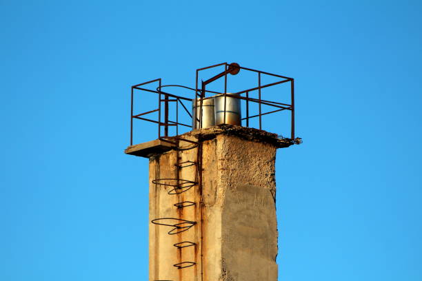 istock Concrete dilapidated cracked industrial chimney with mounted rusted metal steps on one side and fence on top surrounded with clear blue sky in back 1133970581