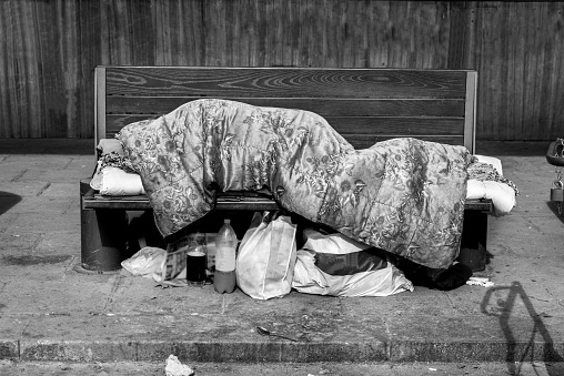 Homeless man, Poor homeless man or refugee sleeping on the wooden bench on the urban street in the city covered with a blanket with bags of clothes and junk on sunny cold day, social documentary concept
