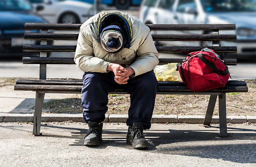 Homeless man, Old poor homeless man or refugee sleeping on the wooden bench on the urban street in the city with bags of clothes on sunny cold day, social documentary concept