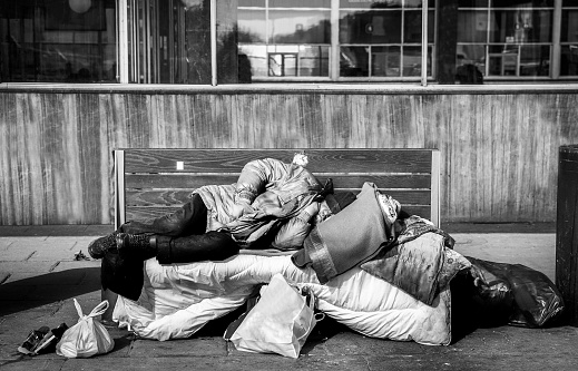 Homeless man, Poor homeless man or refugee sleeping on the wooden bench on the urban street in the city with bags of clothes and junk on sunny cold day, social documentary concept black and white