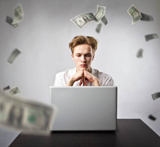 istock Young man using a laptop to browse the net. Young worker and falling dollar banknotes. Currency and job concept. 1133983606