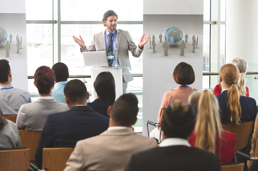 Front view of Caucasian businessman with laptop speaks in front of business colleagues at business seminar in office building