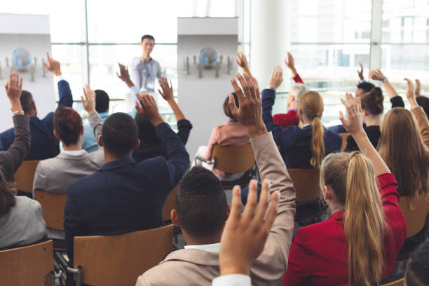 Business people raising their hands at a business seminar Rear view of diverse business people raising hands while they are sitting in front of Asian businessman at business seminar in office building hand raised stock pictures, royalty-free photos & images