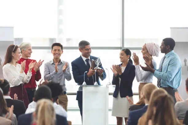 Front view of mixed race businessman holding award on podium with colleagues in front of business professionals at business seminar in office building