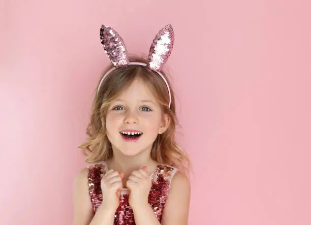 Cute little child wearing sparkling bunny ears on Easter day on pink background. Easter girl portrait, funny emotions, surprise. Copyspace for text.