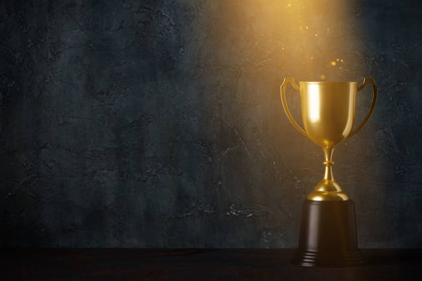 Golden trophy cup on dark table with rays of light Golden trophy cup on dark table with rays of light and glitter dust dark background with copy space gold trophy stock pictures, royalty-free photos & images