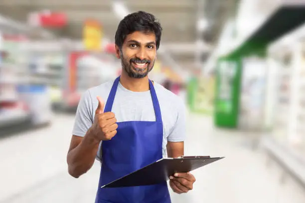 Indian male worker at hypermarket or supermarket showing thumb-up like gesture while holding clipboard