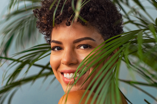 Beautiful young woman with nude makeup looking trought palm leaves. Brazilian smiling woman looking at camera with palm leaves near face. Closeup face of happy african girl in the nature.
