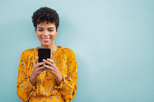 Happy smiling black woman using smart phone. Young brazilian woman writing a message with mobile isolated on blue background. Portrait of african american stylish girl using cellphone with copy space.