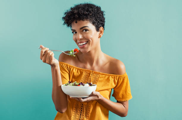 Woman eating salad Beautiful young african woman eating fresh salad with cherry tomatoes. Portrait of happy smiling woman in diet isolated on blue background. Cheerful brazilian girl with curly hair eating fresh vegetable while looking at camera with copy space. mouth full stock pictures, royalty-free photos & images