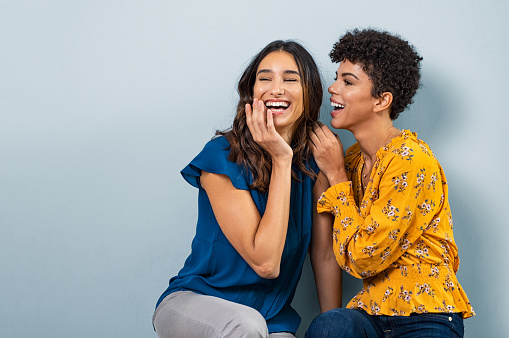 Two best friend girls whispering a secret. Beautiful latin woman and brazilian girl laughing and sharing gossip while sitting against wall with copy space. Stylish friends talking to each other while smiling.