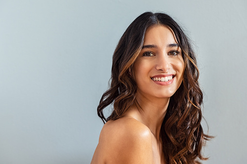 Smiling young woman looking at camera isolated on gray background. Beauty portrait of brunette girl with long curly hair looking at camera. Skin care treatmet and haircare concept at beauty spa.