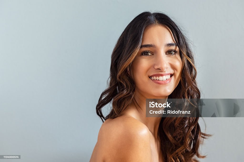 Natural hispanic beauty Smiling young woman looking at camera isolated on gray background. Beauty portrait of brunette girl with long curly hair looking at camera. Skin care treatmet and haircare concept at beauty spa. Women Stock Photo