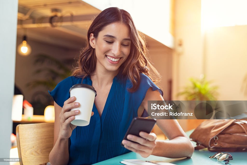 Woman using smartphone Young beautiful woman holding coffee paper cup and looking at smartphone while sitting at cafeteria. Happy university student using mobile phone. Businesswoman in casual clothes drinking coffee, smiling and using smartphone indoor. Women Stock Photo