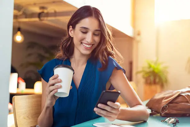 Young beautiful woman holding coffee paper cup and looking at smartphone while sitting at cafeteria. Happy university student using mobile phone. Businesswoman in casual clothes drinking coffee, smiling and using smartphone indoor.