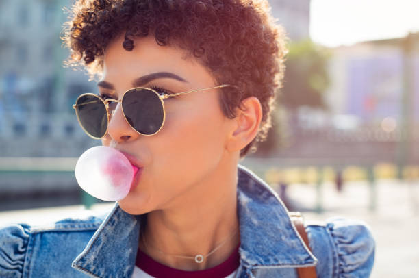 Woman blowing bubble gum balloon Closeup face of african young woman with curly hair blowing bubblegum outdoor. Brazilian girl wearing sunglasses and denim jacket making bubble using chewing gum. Beautiful teenager blowing pink bubble gum. bubble gum photos stock pictures, royalty-free photos & images