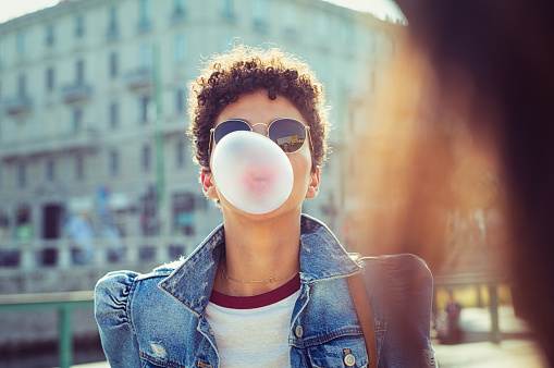 Young african girl in sunglasses with curly hair puffs bubble of chewing gum in city street. Brazilian young woman inflates pink chewing gum. Stylish woman making bubblegum of chewing gum.