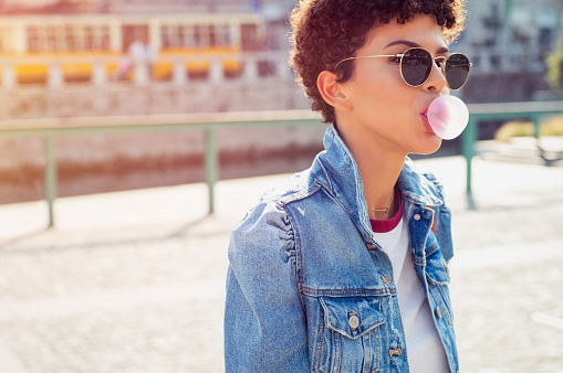 Young beautiful african woman in sunglasses with curly hair puffs bubble of chewing gum in summer. Brazilian young woman inflates pink chewing gum. Stylish woman making bubblegum of chewing gum.